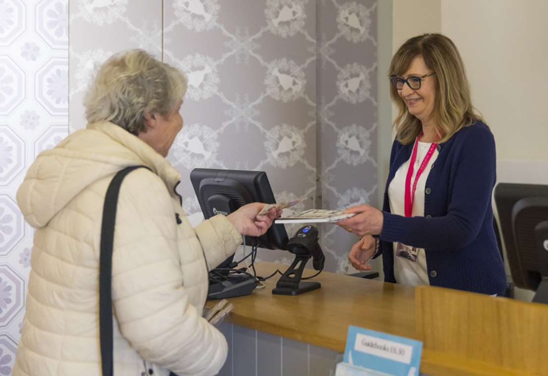 A woman wearing a cream coloured coat and handbag has short grey hair and is in her late 60s, handing over a £10 note to a staff member at a till.