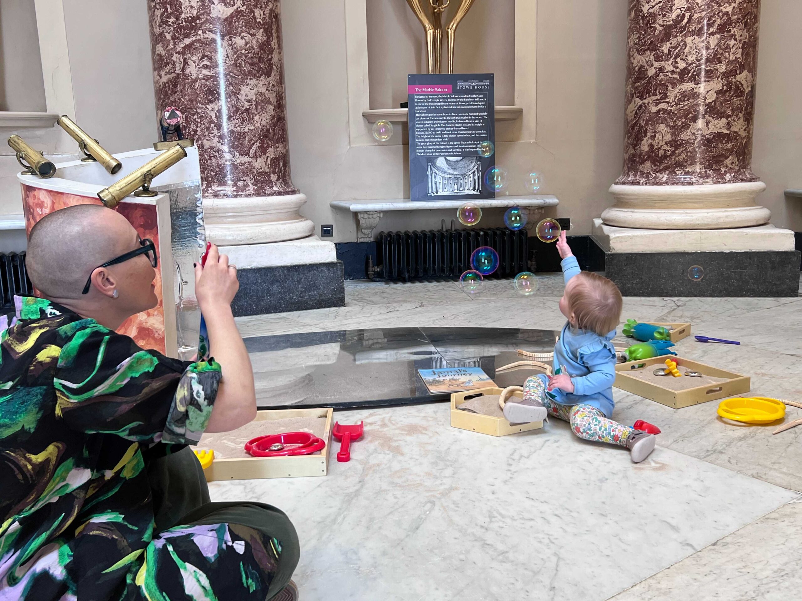 A baby girl approx age 1 and a half sits and stretches for some bubbles floating through the air which have been blown by a session leader sitting on the floor. Toys are scattered around for young toddlers to play with. The room is a large room with Roman style columns