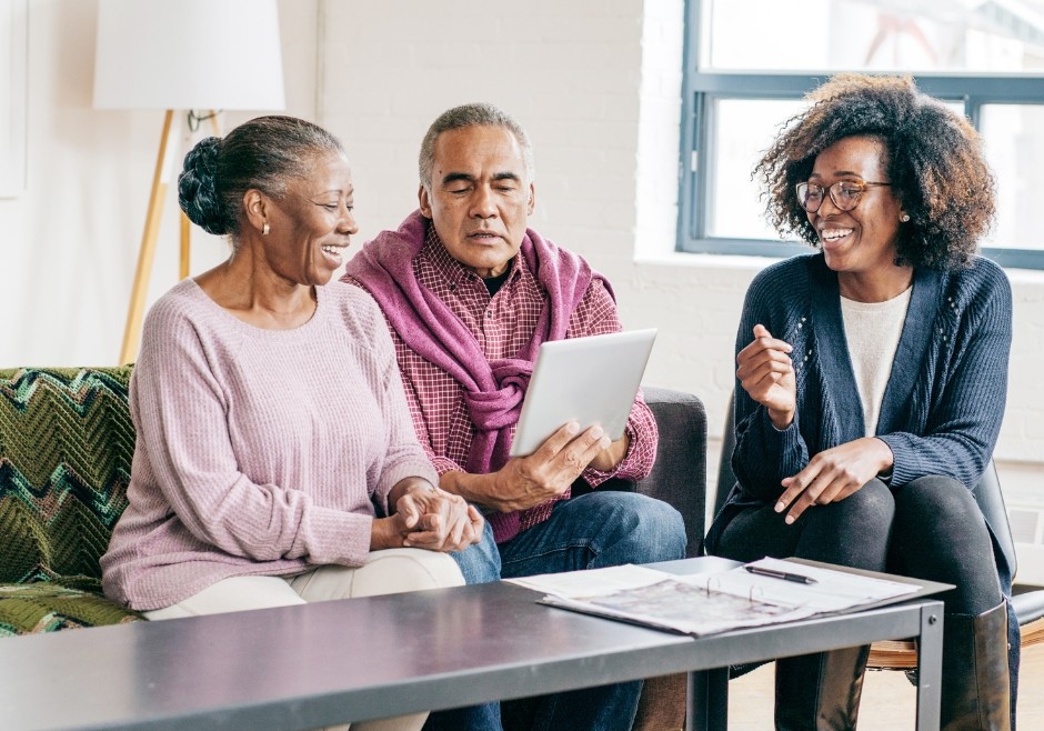 A husband and wife sit together on a sofa, they are black and in their late 60s. One is holding a document which they both look at and smile. A lady sits in a chair to the right, also looking towards the document. they are updating their wills.