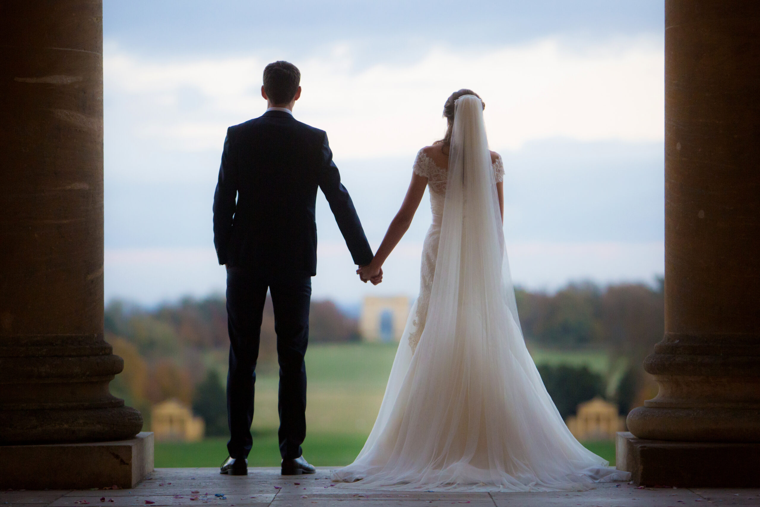 A man and woman stand on between two Roman columns and look out towards the landscape garden at Stowe. They are a bride and groom in traditional black suit and white long dress with veil.