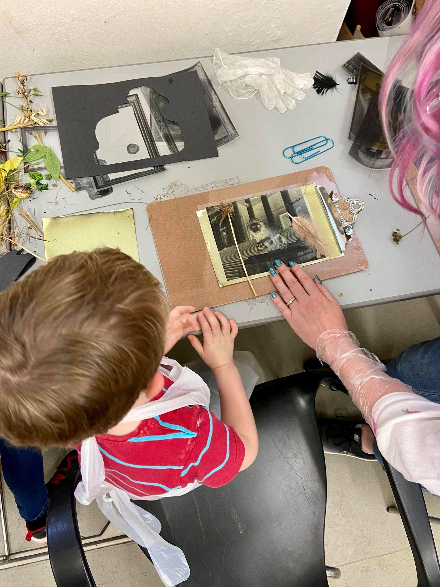 A youg boy with dark blonde hair and red tshirt wears an apron and looks at his artwork. There is a mother helping him with the cyanotyping activity