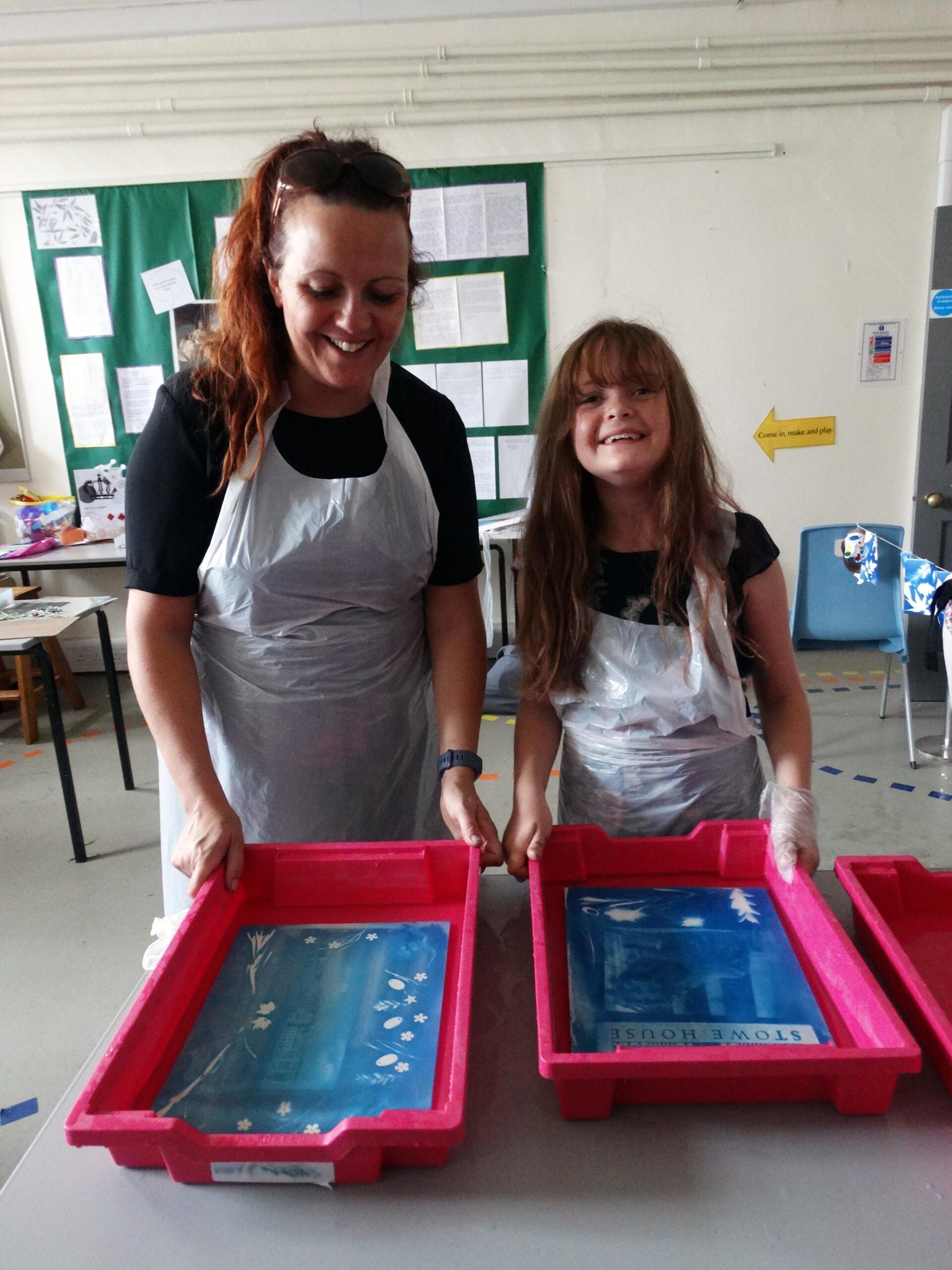 A mother in her 30s and daughter approx 10 hold a tray with the designs from the cyanotype workshop. They both smile and have long brown hair.