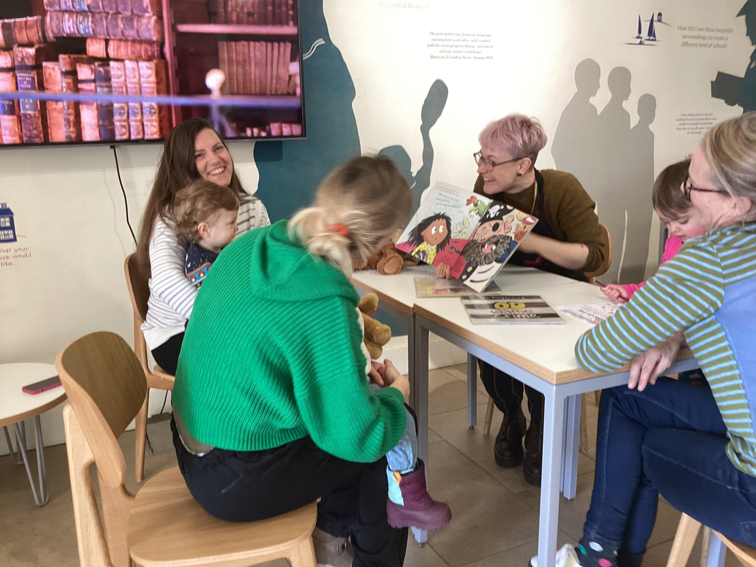 A woman with short hair smiles and holds a book as she sits at a table and reads stories to babies and toddlers who are sitting on their mother's laps.