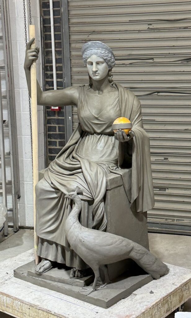 A seated clay statue of a goddess holding an orb and wooden stick with a peacock next to her feet.