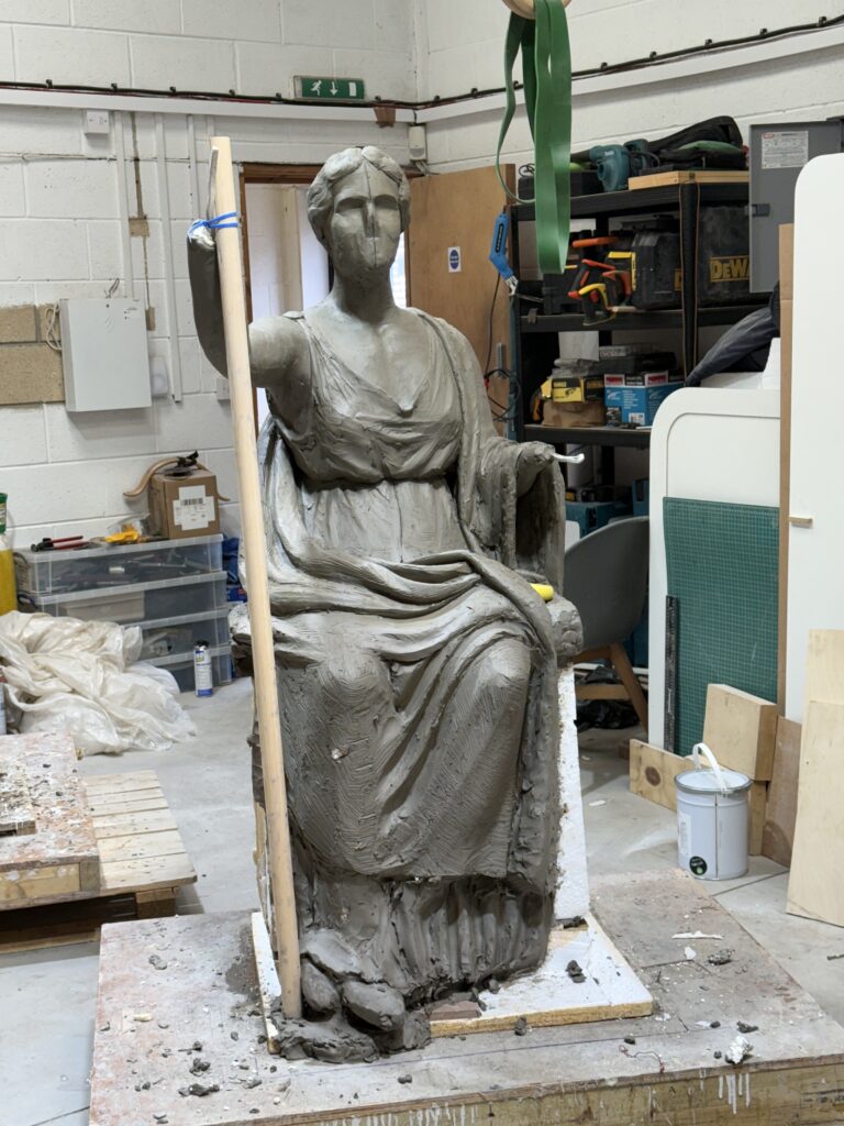 A clay model of a seated goddess holding a wooden stick