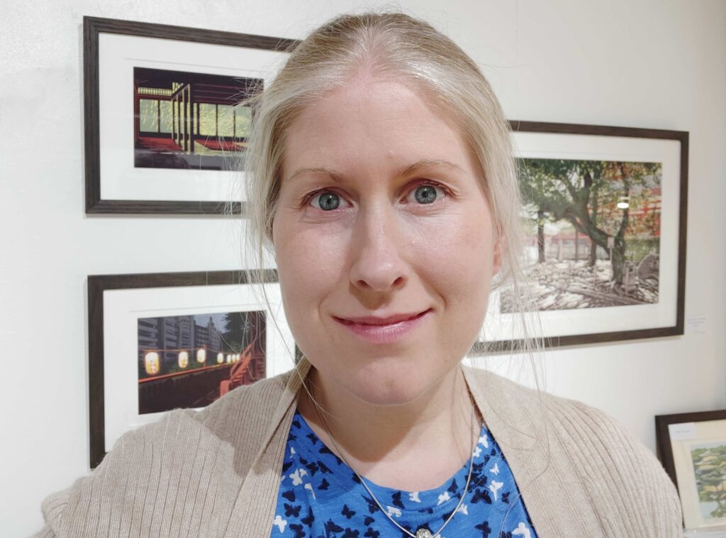 a white lady in her 40s with a light complexion and pink lips has a closed lip smile and a soft expression as she looks at the camera. She has blonde hair tied U in a ponytail and stands in front of a wall of her framed artwork. She is the artist Alexandra buckle