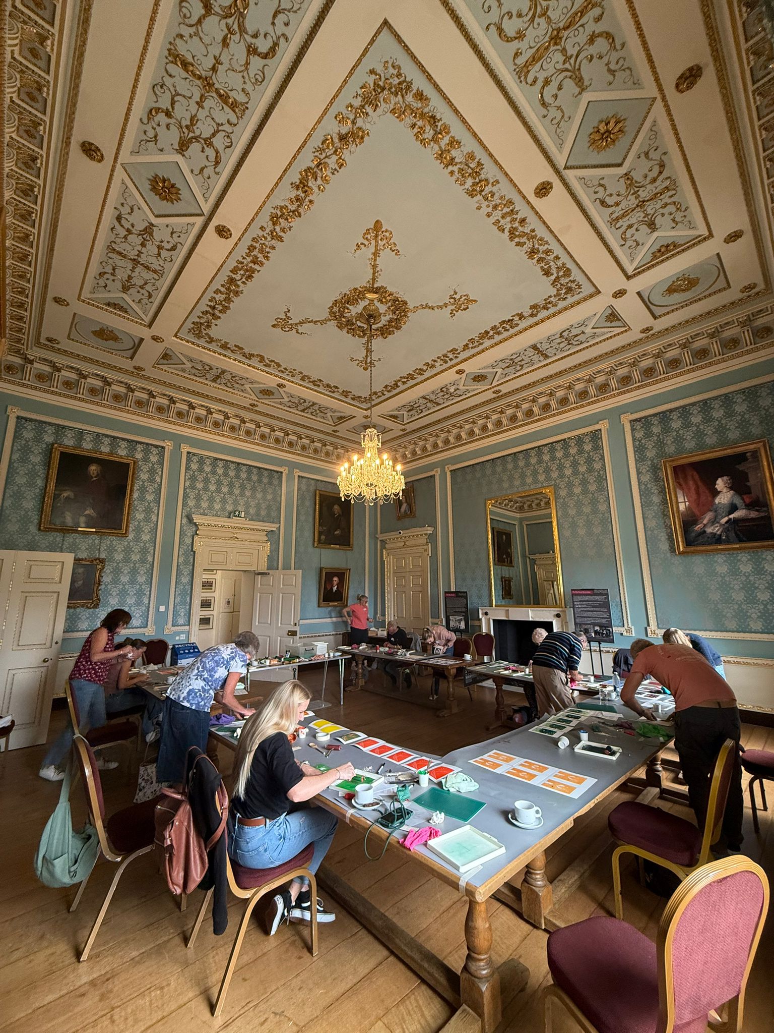 An ornate room with blue wallpaper and carved plaster ceiling which is gilded with gold is set up with a rectangle formation of tables as people sit and partake in a lino print workshop.