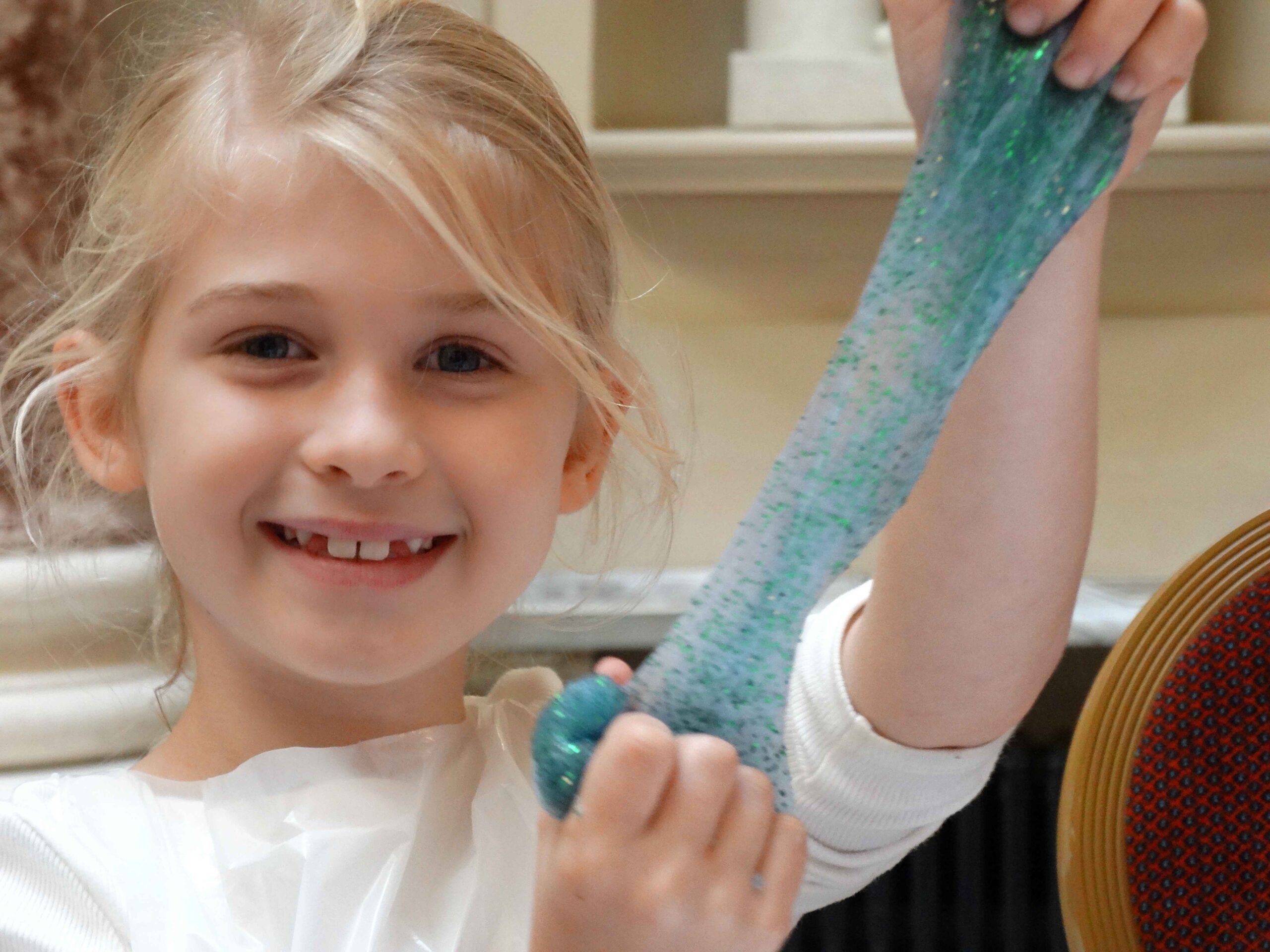 Smiling child stretching a strand of blue-green glittery slime between her hands.