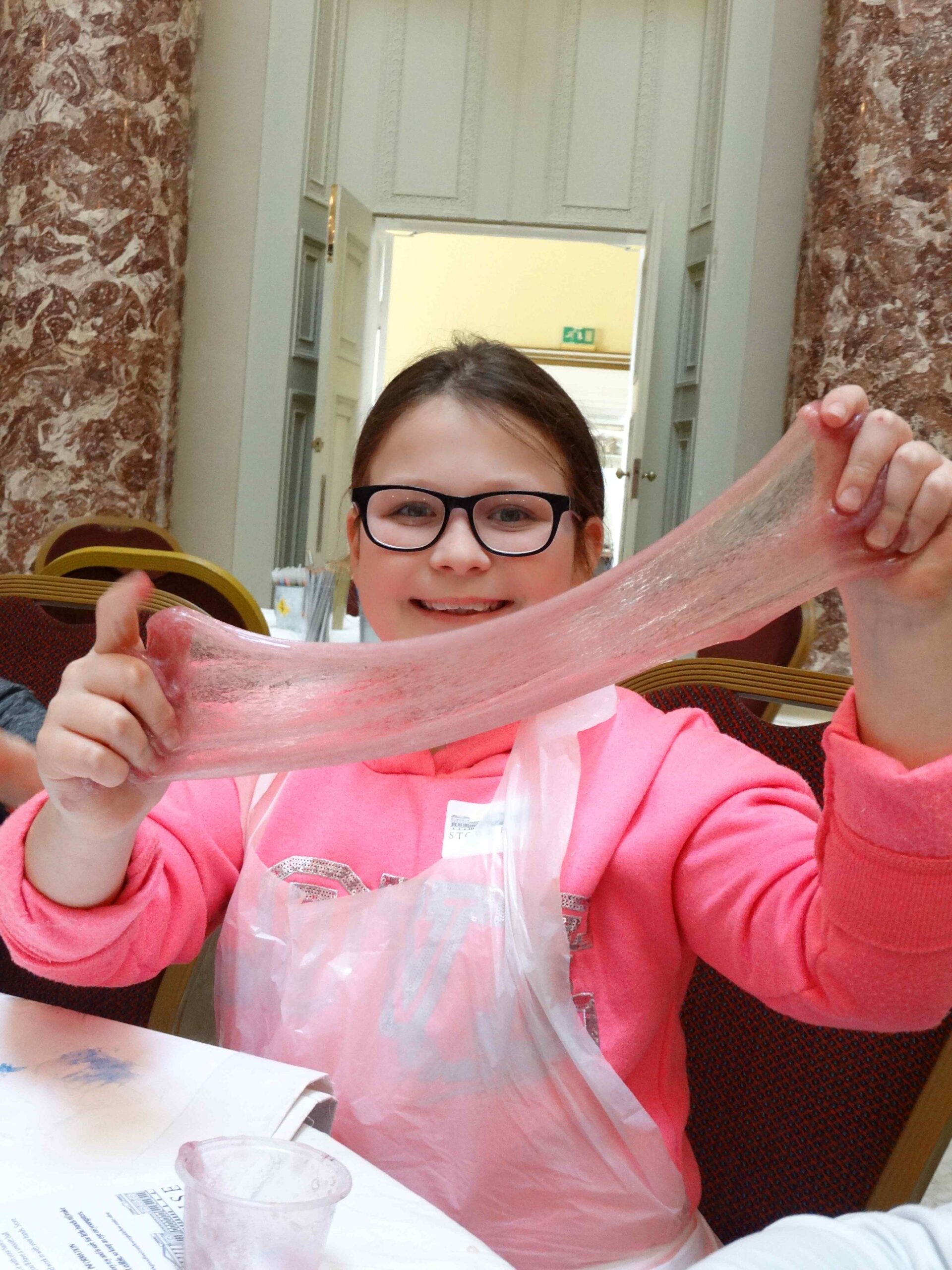 Smiling child wearing glasses and stretching a wide sheet of pale pink slime.