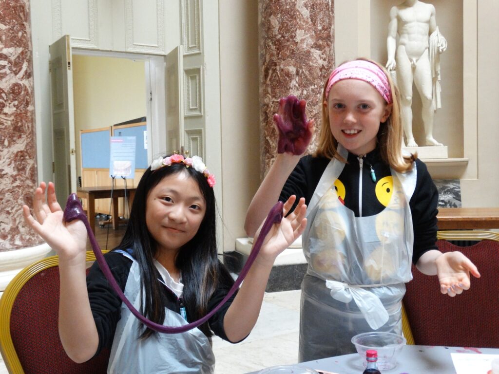 Two children smiling as they stretch and display purple slime during a workshop at Stowe House.
