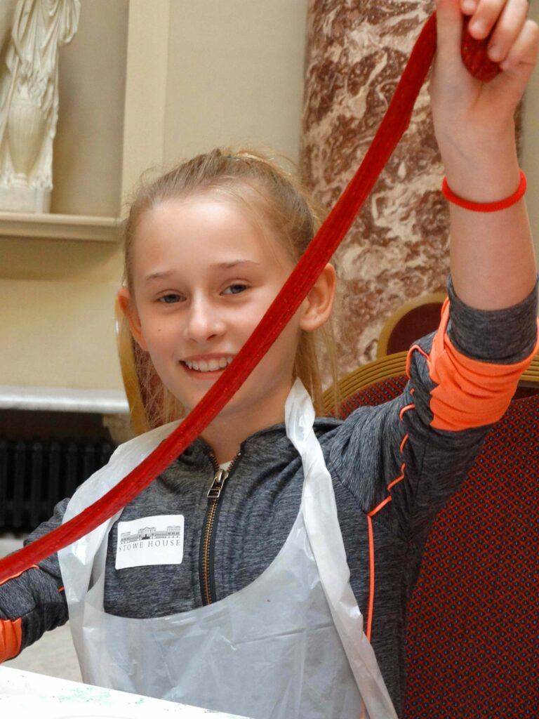 Smiling child stretching a long strand of glittery red slime above their head.
