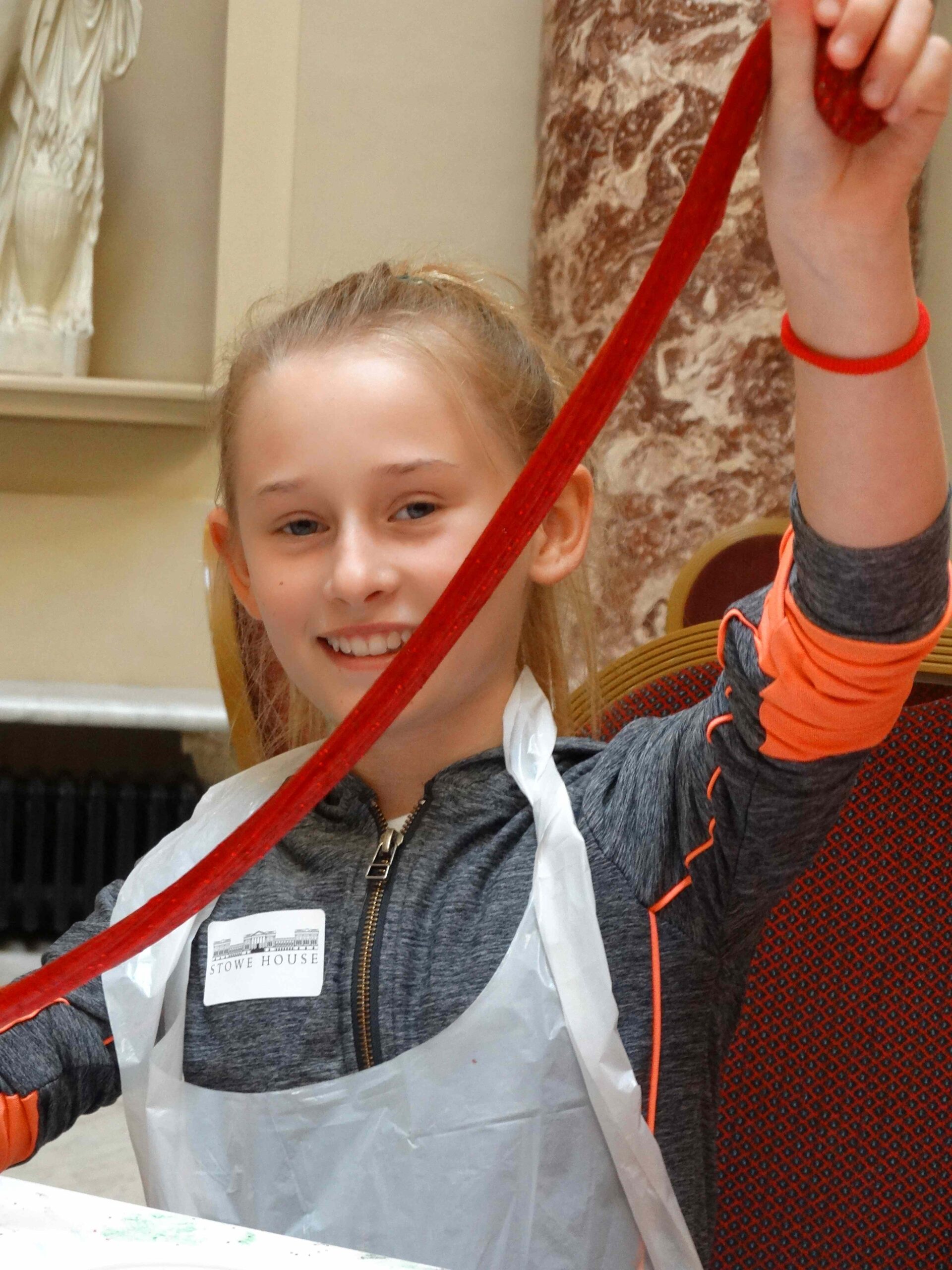 Smiling child stretching a long strand of glittery red slime above their head.