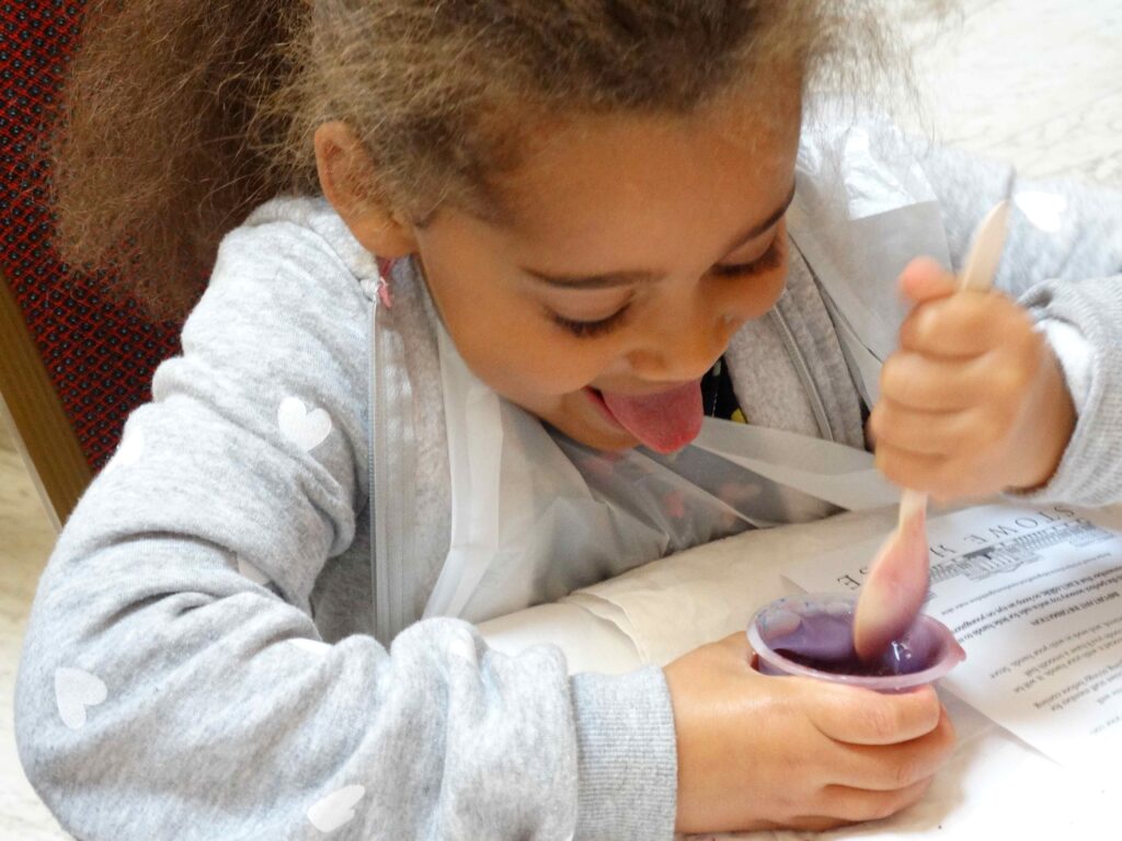 Child playfully sticking out their tongue while mixing purple slime in a small container.